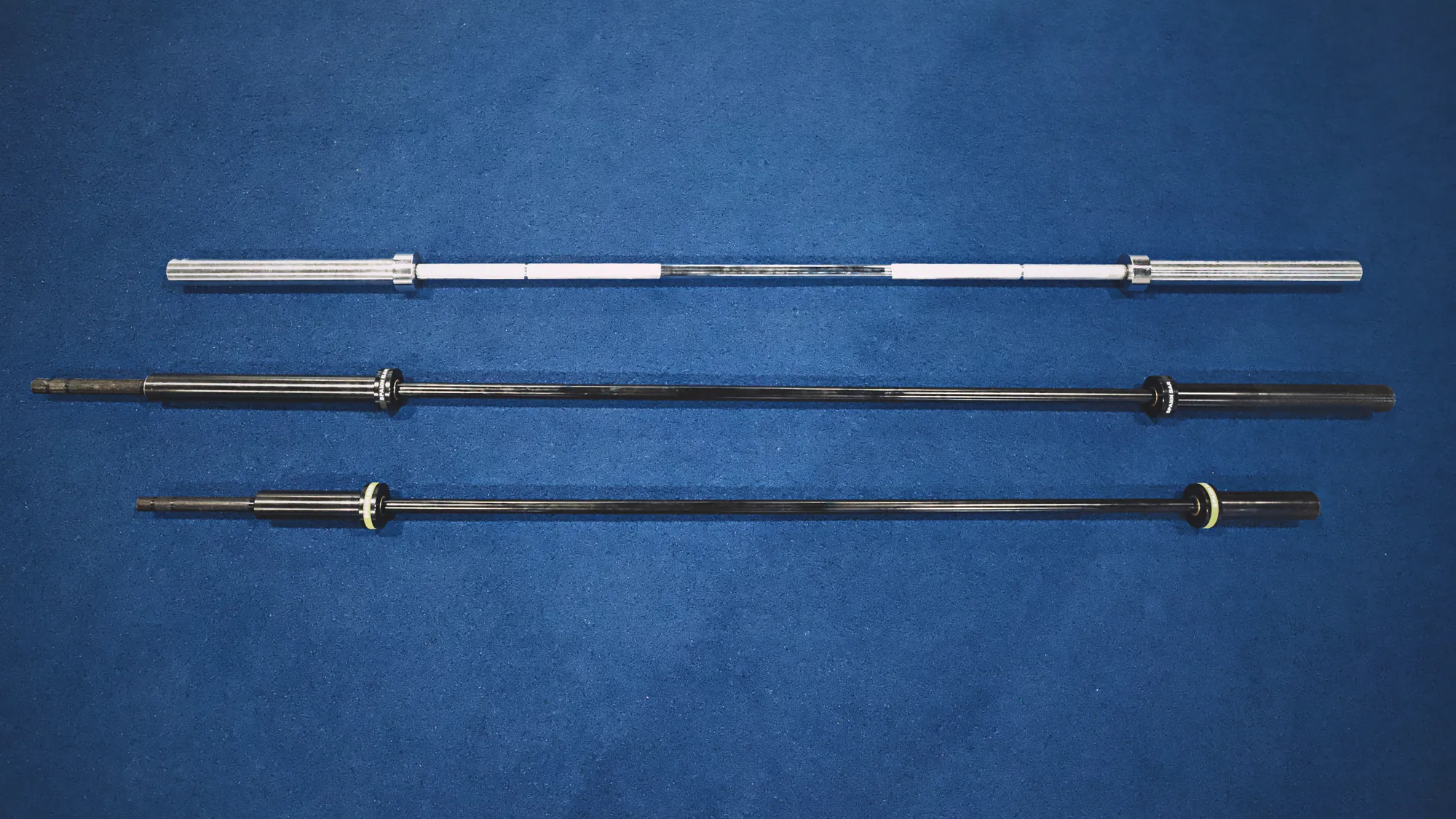 Three bars laid on blue turf: the 43 lb Spann Bar with its black collar, the 25 lb Melo Bar with its yellow collar, and a chrome standard Olympic bar.
