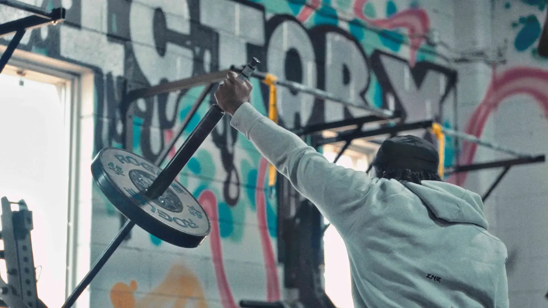 An athlete from behind, pressing the Spann Bar overhead in front of a graffiti wall that reads Victory.