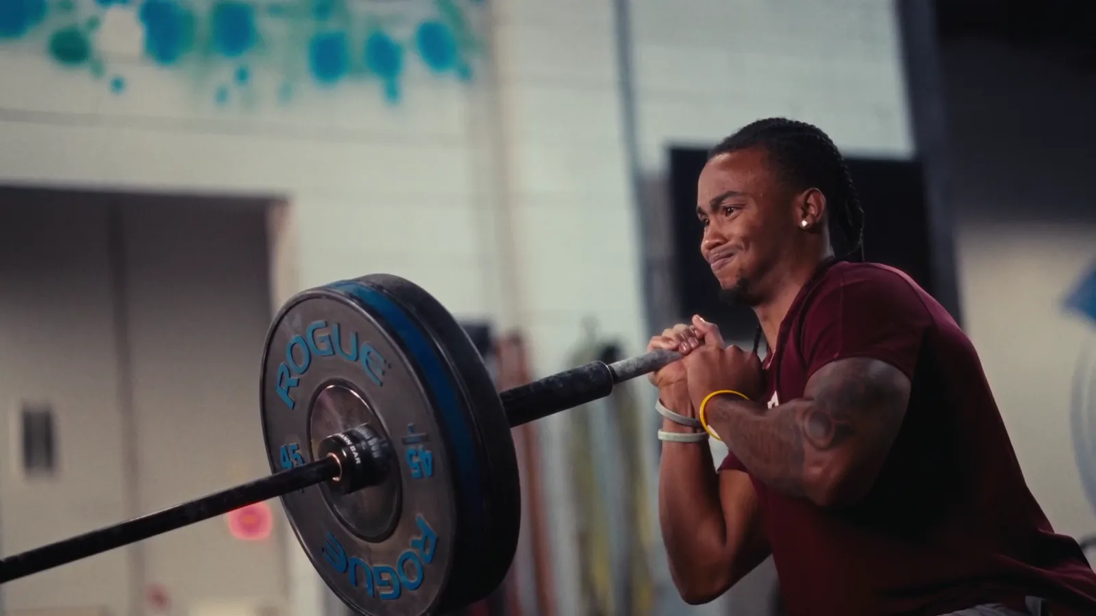 An athlete pressing the Spann Bar overhead one-handed in a landmine press, a bumper plate on the loaded end, a graffiti wall behind.