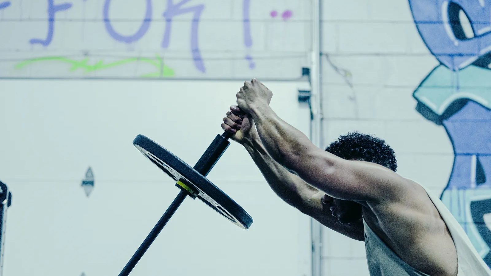 A lifter at the top of a landmine press with the Melo Bar, yellow collar on the sleeve, against the graffiti wall at SPS Fit