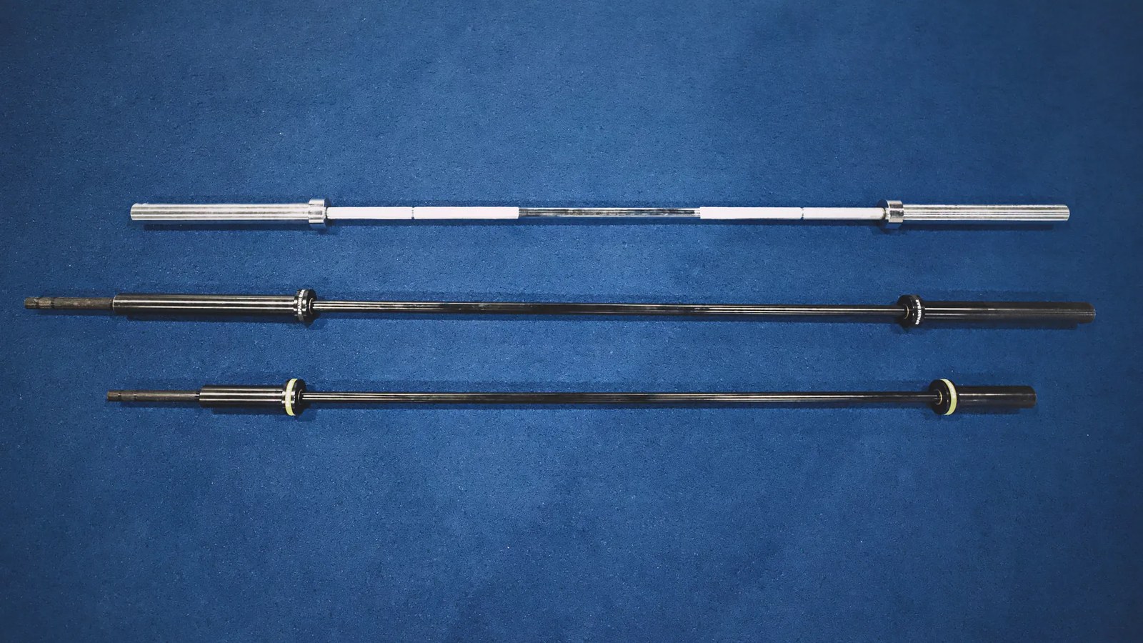 Three bars laid on blue turf at SPS Fit: the 43 lb Spann Bar with its black collar, the 25 lb Melo Bar with its yellow collar, and a chrome standard Olympic bar.